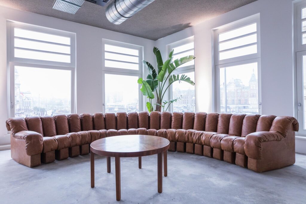 L-vormige bruine leren bank met ronde houten tafel in een lichte ruimte aan de Prins Hendrikkade met uitzicht op Amsterdam Centraal.